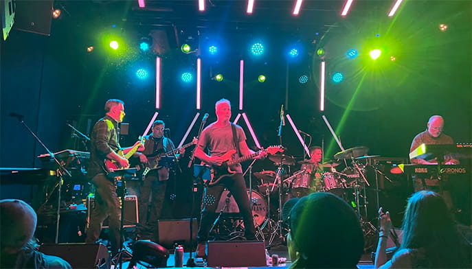 A band performs on stage under colorful lights, with musicians playing guitar, bass, drums, and keyboard while an audience watches.