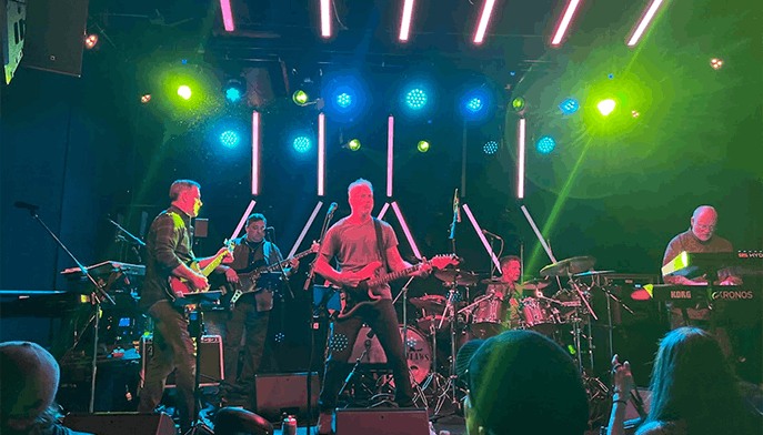A band performs on stage under colorful lights, with musicians playing guitar, bass, drums, and keyboard while an audience watches.