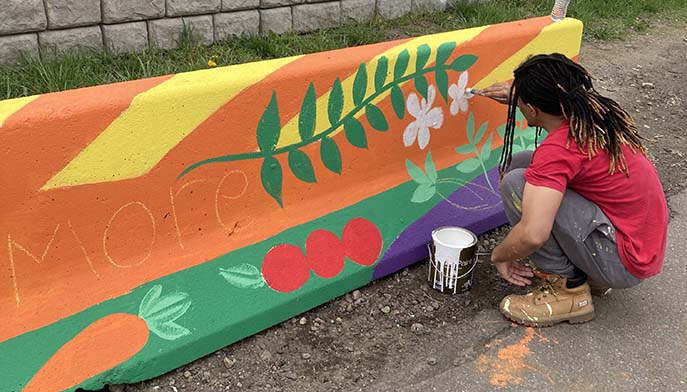 Person kneels and paints a colorful mural with leaves, flowers, and vegetables on a concrete barrier outdoors.