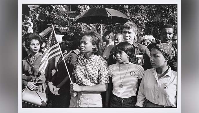 A black and white photograph by Danny Lyon titled “SNCC workers stand outside the funeral: Emma Bell, Dorie Ladner, Dona Richards, Sam Shirah, and Doris Derby, Birmingham” depicting a group of solemn young African American individuals, some holding American flags, standing closely together waiting outside a funeral.