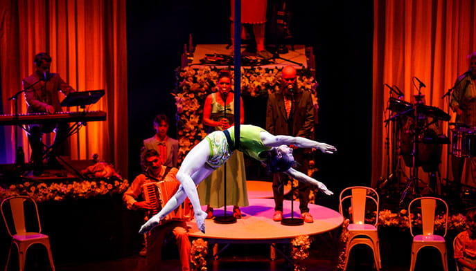 An aerialist performer hangs in the air from a ribbon wrapped around their waist while a singer and band perform.