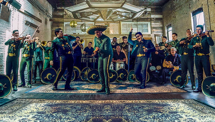 Fourteen-member Latin mariachi band in matching attire performing one of their hits, complete with violins, trumpets, trombones, drums, and an iconic sombrero hat.