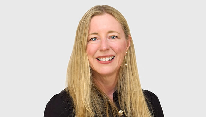 Shannon Harding, PhD, with long blonde hair smiles, wearing a black top and long gold earrings. The background is plain light grey, conveying a professional yet approachable tone.