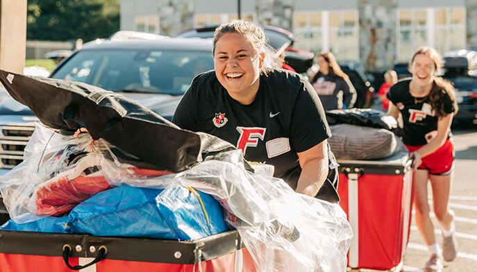Smiling volunteer in a black F-shirt pushes boxes in a cart as others push red bins outdoors in bright sunlight.