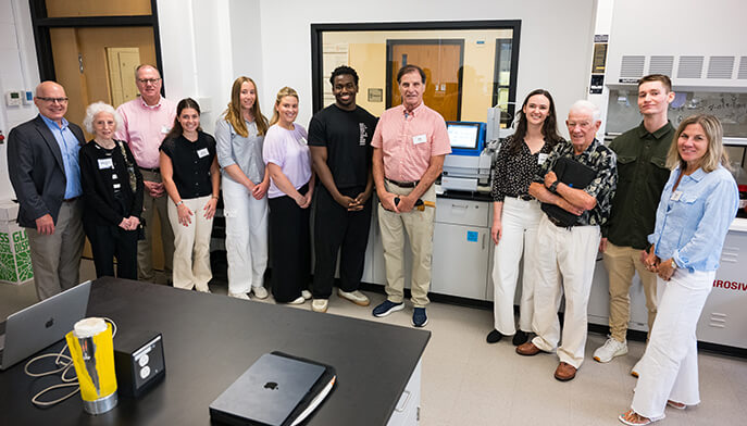 Diverse group of 12 adults poses in a laboratory beside medical equipment, smiling warmly and conveying teamwork and community.