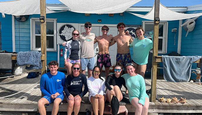 Smiling group of ten students pose on a sunny waterfront dock beside a blue beach house, wearing casual summer clothes and swimwear.