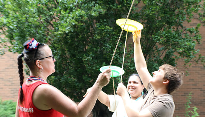 Three people outdoors at Fairfield University’s Flag Day Celebration joyfully practice spinning colorful plates on sticks at the Circus Moves station, smiling and laughing amid leafy trees and a brick building.