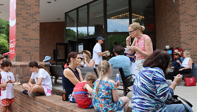 Families and friends gather outside the Regina A. Quick Center for the Arts at Fairfield University’s Flag Day Celebration, eating and chatting with drinks; a lively, relaxed community scene unfolds on the patio area in front of the brick building.