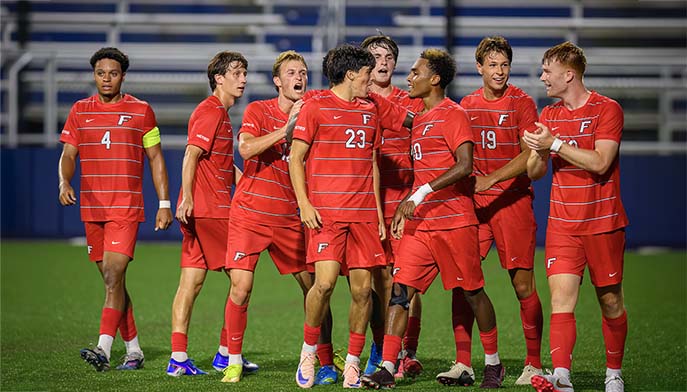 Players from the men's soccer team celebrate a goal, hugging and high-fiving in a moment of joy and camaraderie.