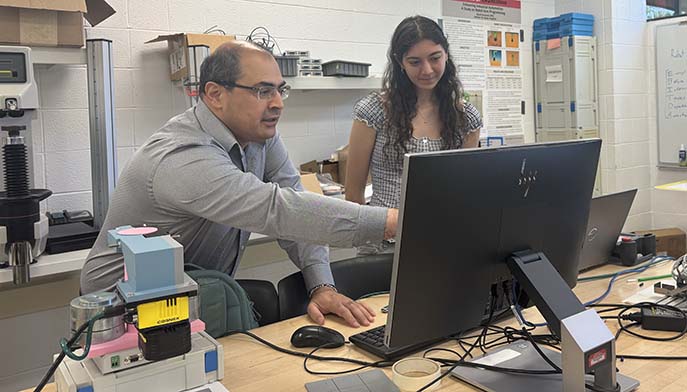 Two people work at a computer workstation in a lab, with one pointing at the monitor and lab equipment on the table.