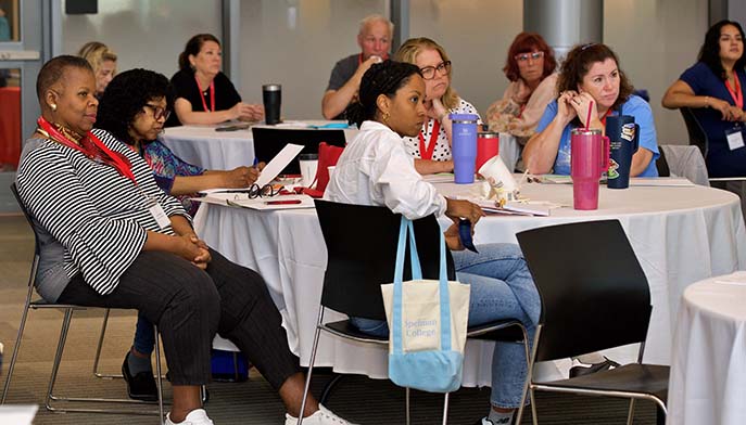 Attendees seated at round tables during a workshop or conference session, listening and taking notes.