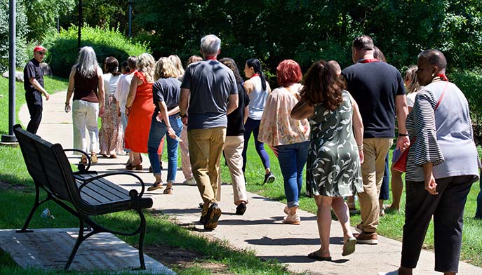 Group of adults walking together along a sunlit park path, with a bench in the foreground and trees and grass around them.