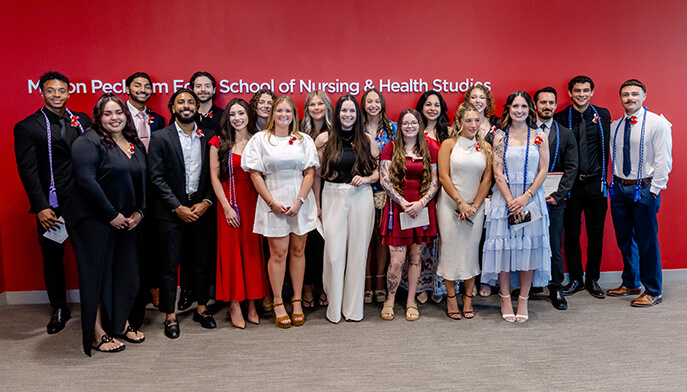 Group photo of diverse nursing and health studies graduates posing in formal attire before a red backdrop, smiling in a celebratory mood.