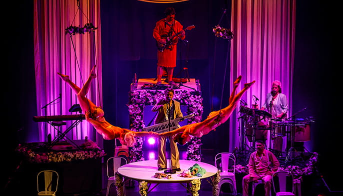 Colorful theatrical stage bathed in purple light, with musicians, a central performer, and two orange-clad acrobats forming a dramatic arch.