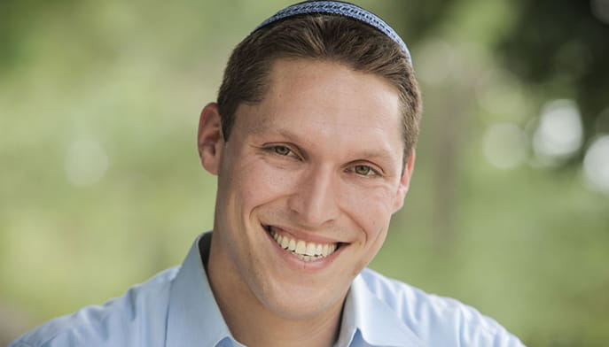 Smiling Rabbi Shmuly Yanklowitz wearing a blue kippah and light blue shirt, posed outdoors against a softly blurred green background; warm, friendly mood.