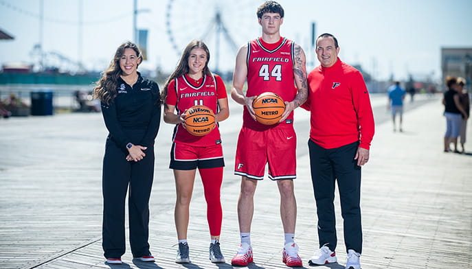 One graduate and one sophomore student-athlete for Fairfield's Women's and Men's Basketball teams (respectively) posing with basketballs and their coaches on a sunny boardwalk.