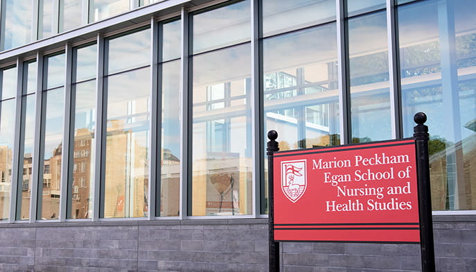 A close-up of the red, branded sign that reads 'Marion Peckham Egan School of Nursing and Health Studies', marking the modern, brick-and-glass building.