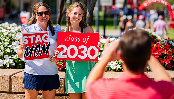 A first-year student holding up a written sign for 'Class of 2030' with her mom (with a written sign for 'Stag Mom') being photographed at move-in.