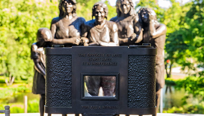 Photograph taken behind the memorial sculpture's bronze camera, which reminds the viewer with an inscription as they look upon the six immortalized, metal women, ‘The opposite of love is not hate—it is indifference. You are a witness’.