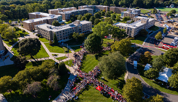 Aerial view of the Fairfield University campus as a large, colorful crowd gathers and marches along a central lawn, creating a lively community scene.