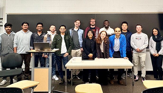 A diverse group of students and instructors poses behind a classroom table and laptop, smiling in a relaxed, welcoming atmosphere.