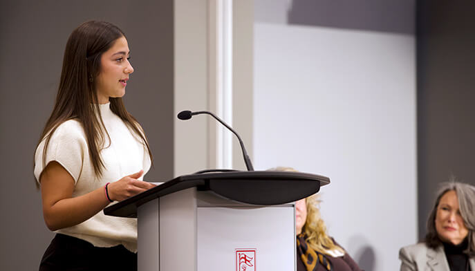 Isabella Morales speaks confidently at a lectern with a microphone, while two seated women watch in a bright presentation room.