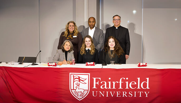 Six people pose behind a table draped with a red Fairfield University banner, smiling at a campus event in a bright, welcoming room.