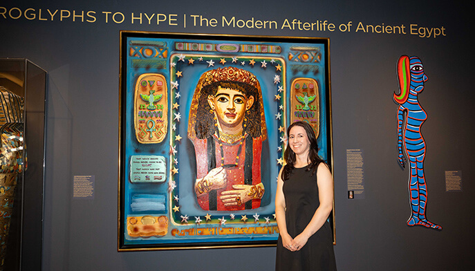 A woman in a black dress, curator Megan Paqua, poses beside a colorful Egyptian-inspired portrait, Audrey Flack’s “Isis” (1983), at “Hieroglyphs to Hype: The Modern Afterlife of Ancient Egypt,” an exhibition at the Fairfield University Art Museum.