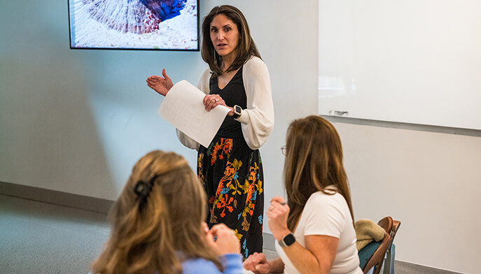 Dr. Kimberly Doughty presents to a small seated audience, holding papers and gesturing beneath a wall-mounted screen; the attentive discussion feels informal and focused.