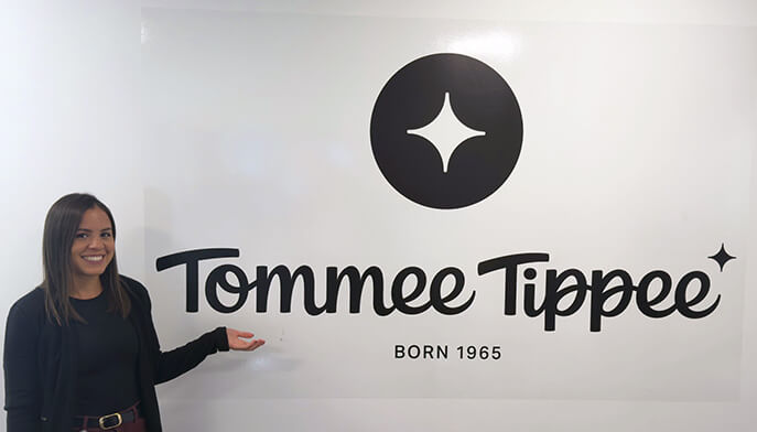 A smiling Mayana Dos Reis MBA’26, MS’26 gestures toward the Tommee Tippee logo on a white wall, with its black star emblem and “Born 1965” text; the scene feels welcoming.