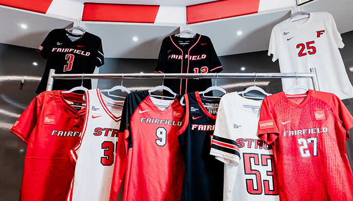 A display of Fairfield University sports jerseys on racks, featuring red, black, and white basketball and team shirts with bold numbers and logos.