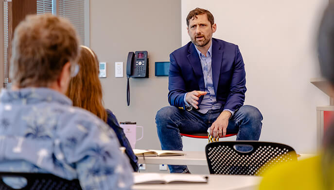 Phil Klay in a navy blazer sits on a table, speaking to a small group in a bright meeting room; listeners face him attentively.