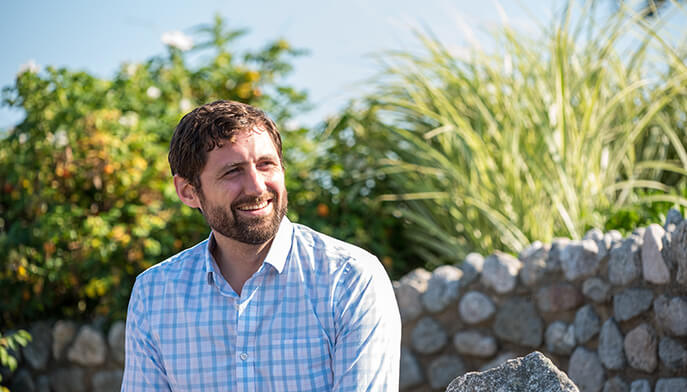 Phil Klay in a blue-and-white checked shirt sits outdoors by a stone wall, with lush greenery behind him in warm sunlight.