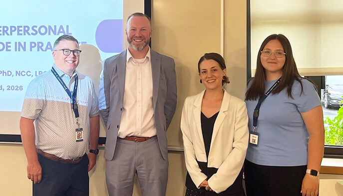 Four smiling people pose together at a professional event, with a presentation screen behind them, conveying a warm, collaborative atmosphere.