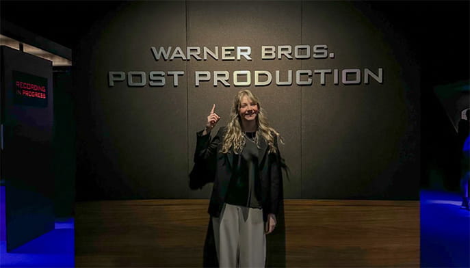 A smiling woman points to the “Warner Bros. Post Production” sign on a dark studio wall, standing in a dim, modern hallway with blue-lit accents.