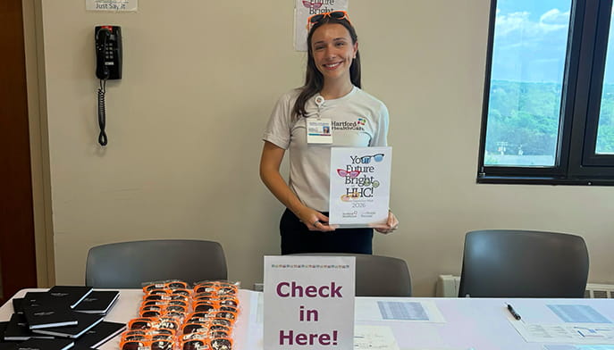 Smiling woman in a white shirt holds a “Your Future Is Bright” certificate behind a “Check in Here!” table lined with brochures and orange items.