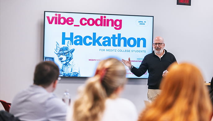 A smiling instructor presents a “vibe_coding Hackathon” screen to seated college students in a classroom, creating an upbeat, collaborative learning mood.