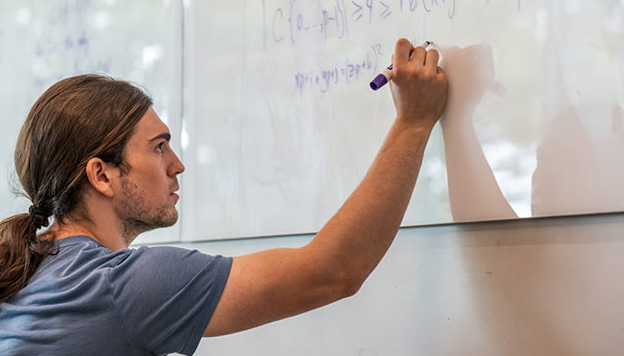 A focused young man with long hair writes equations on a glass whiteboard with a purple marker in a bright classroom, conveying concentration.