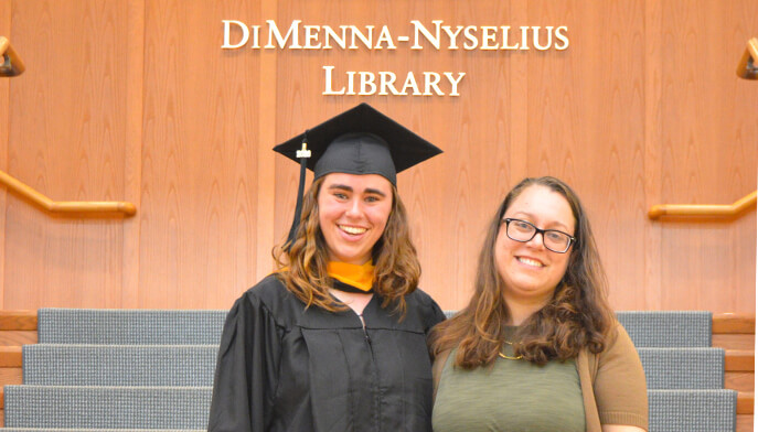 Mia Swenson ’26 in cap and gown poses with head of library beneath the DiMenna-Nyselius Library sign, sharing a warm, proud moment.