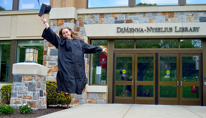 Recent Fairfield Meditz alumna Mia Swenson ’26 in a black gown leaps with one arm raised outside DiMenna-Nyselius Library, celebrating graduation with joyful energy.