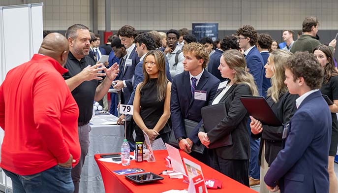 A group of people gathered around a table, attentively listening to a man speaking to them.