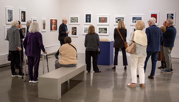 Visitors gather in a bright contemporary art gallery, viewing framed works in the exhibition “James Welling: Cento” as a man, artist James Welling, speaks; the calm, social atmosphere suggests an exhibition tour.