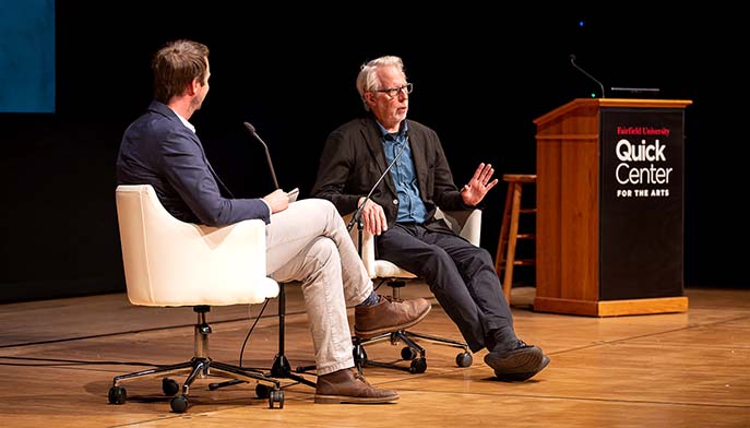 Two men, Andrew Ward and James Welling, converse onstage in white chairs beside a podium labeled “Quick Center for the Arts.” The older speaker, Welling, gestures, creating a thoughtful, formal interview atmosphere.