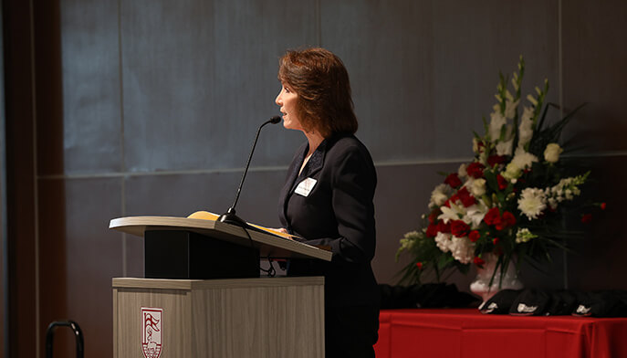 Dean Boyce in a black suit speaks at a lectern with an open book, facing an audience off-frame; a colorful floral arrangement adds a formal, dignified tone.