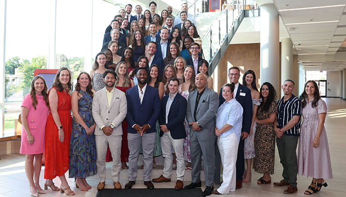 A diverse group of students pose smiling in a bright office atrium beside a staircase, conveying a welcoming, celebratory mood.