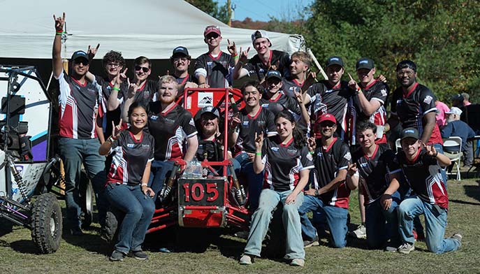 A group of enthusiastic students posing together at an outdoor event, wearing matching team shirts and showcasing their vehicle number 105.