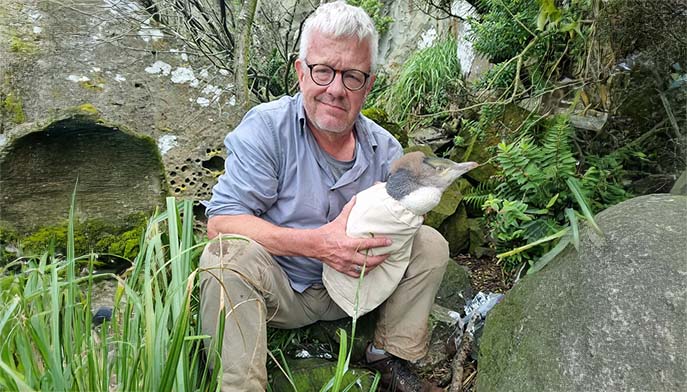 A man in glasses holds a wrapped bird while sitting on a rock amidst lush greenery.