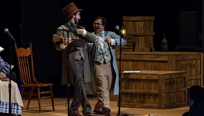 Two actors in period costumes perform on stage beside a microphone and stacked wooden shipping crates.