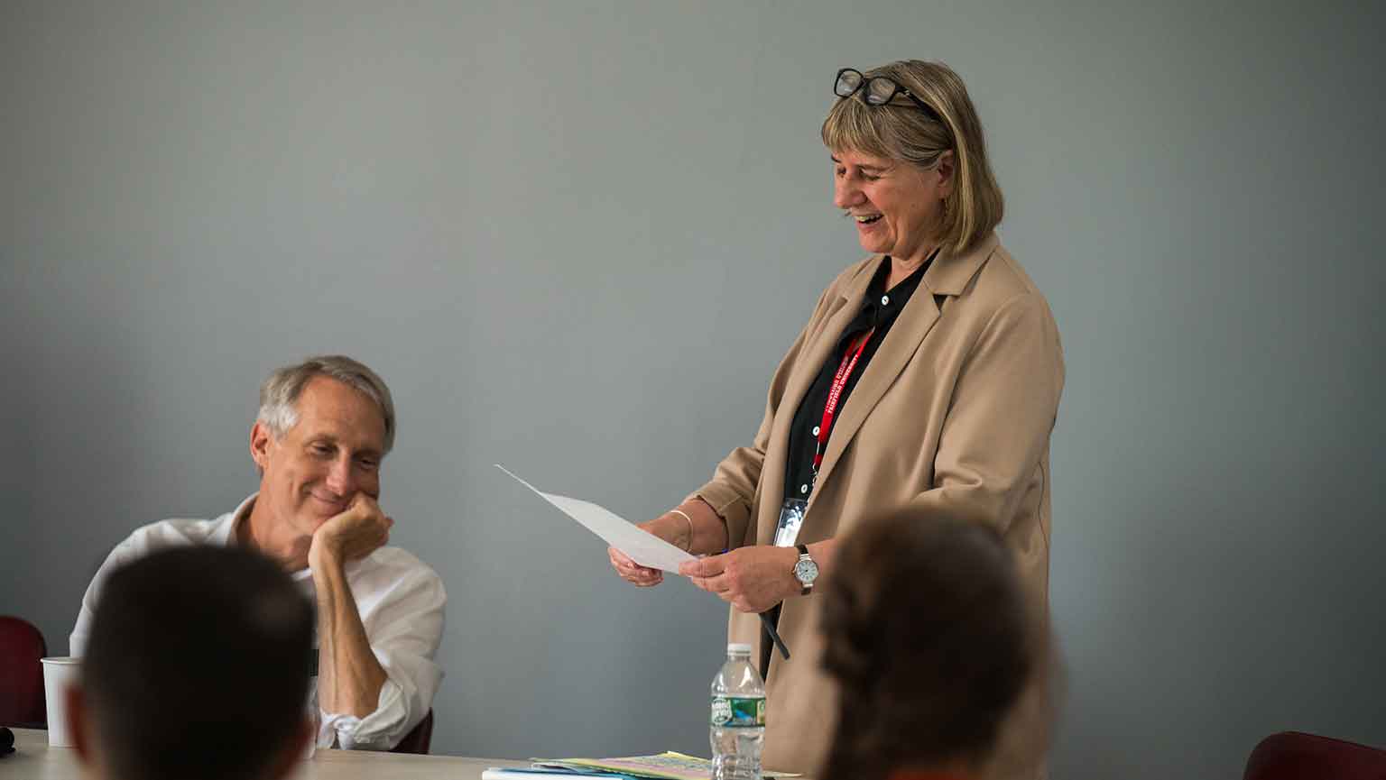 Woman standing at table speaking while people are seated listening.