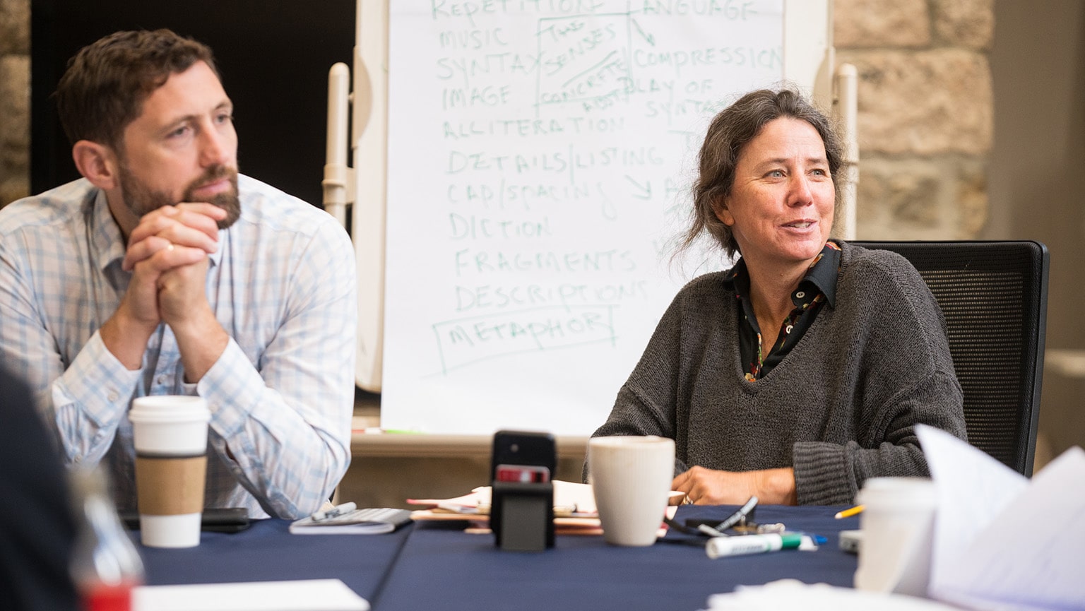 Two MFA faculty seated at a table, engaged in discussion with a whiteboard in the background at a writers workshop.